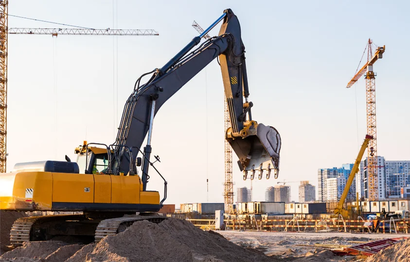 A yellow excavator lifts dirt at a busy construction site, with cranes and partially built structures in the background, representing heavy machinery.